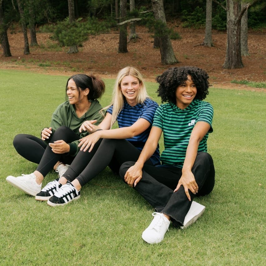 Three women sitting on grass among tall pines, relaxed and talking