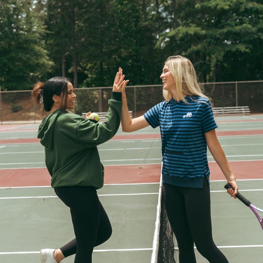 Two women meeting for a high five across a tennis net, both in Moody Bobby tops