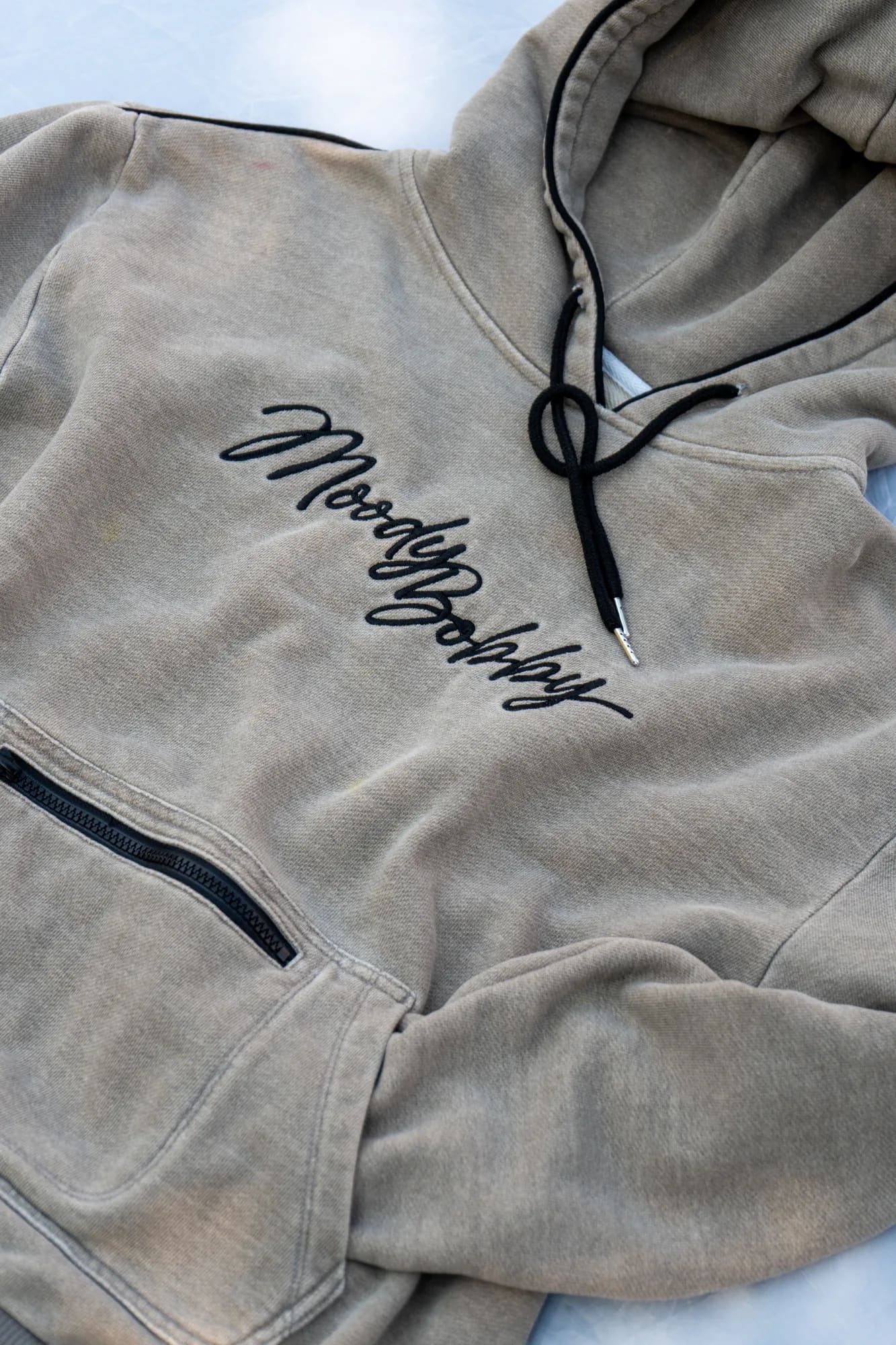 An oatmeal Moody Bobby hoodie laid flat, the embroidered script and the zipper pocket in frame