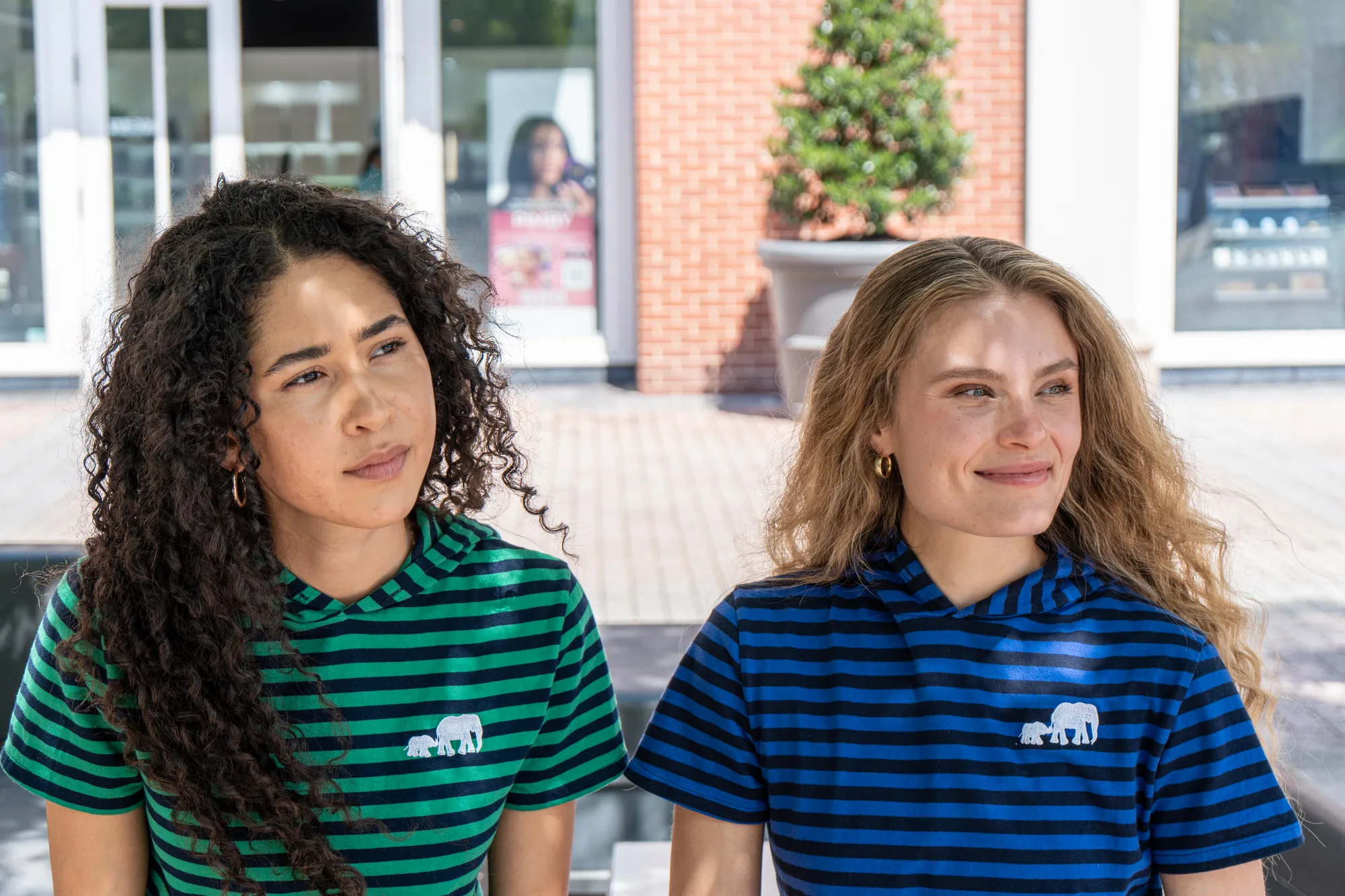 Two women seated side by side outside a storefront, in green and blue striped Moody Bobby hoodie tees