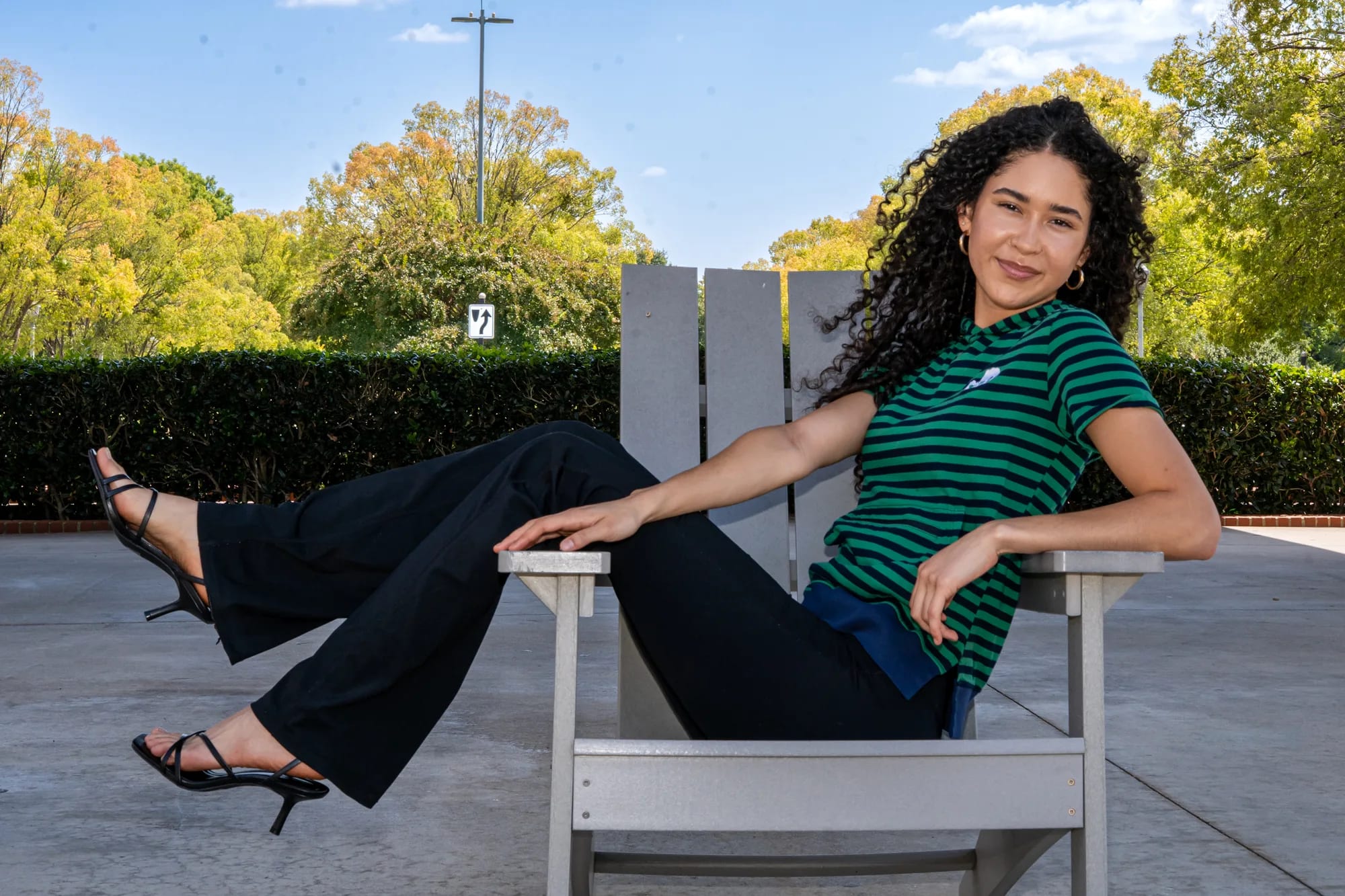 Woman reclining in a white deck chair in a sunlit courtyard, in a green striped Moody Bobby hoodie tee