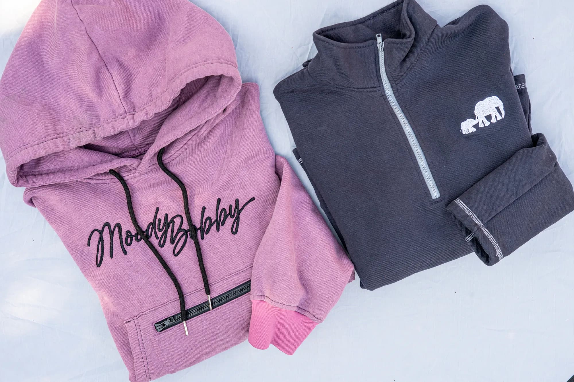 Two folded Moody Bobby hoodies shot from above, a mauve pullover beside a charcoal quarter zip