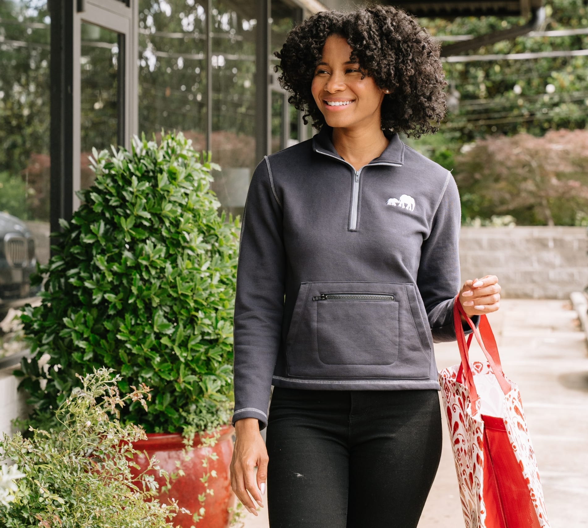 Woman in a grey quarter zip walking through a garden nursery carrying shopping bags