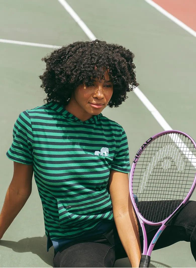 Woman in a green striped Moody Bobby hoodie tee sitting on a tennis court, racket resting against her knee