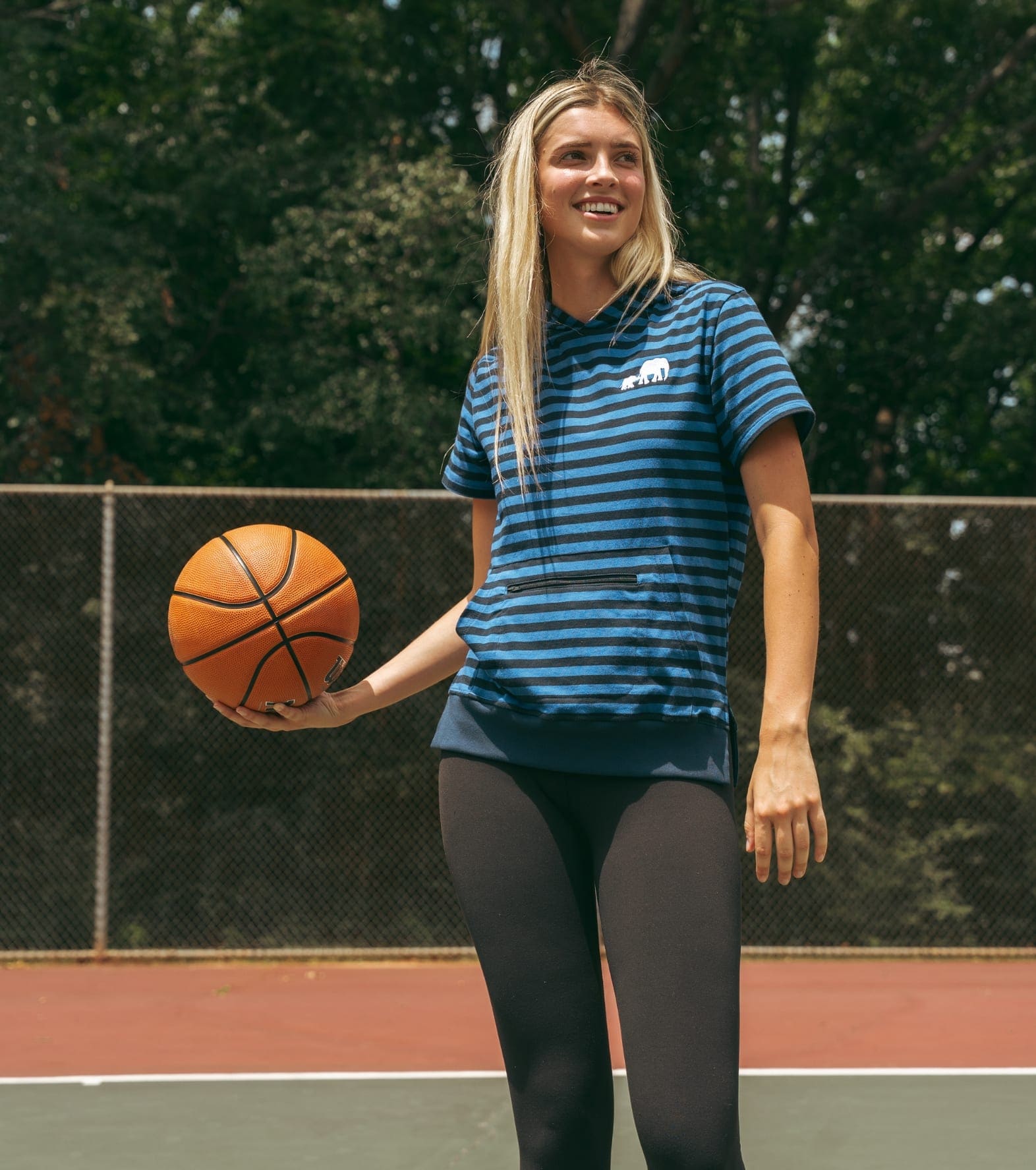 Woman in a blue striped tee standing on an outdoor court, holding a basketball at her side
