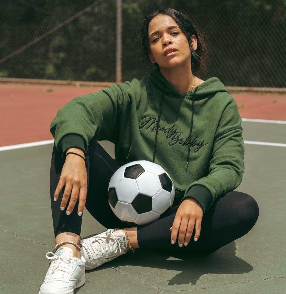 Woman in an olive green hoodie sitting on an outdoor court with a soccer ball resting against her knee