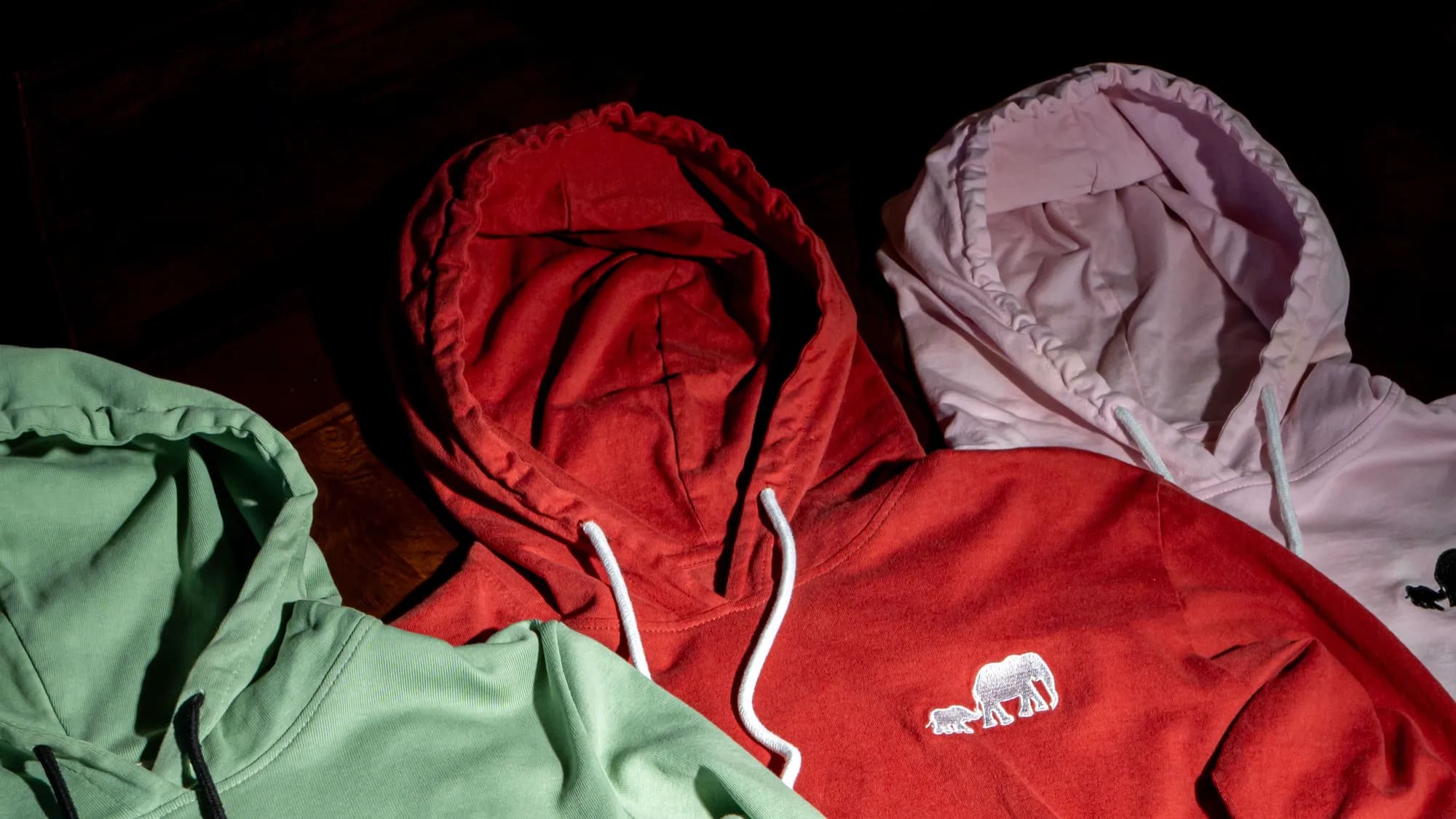 Three hoodies folded together in sage green, deep red and pale pink, photographed on a dark surface