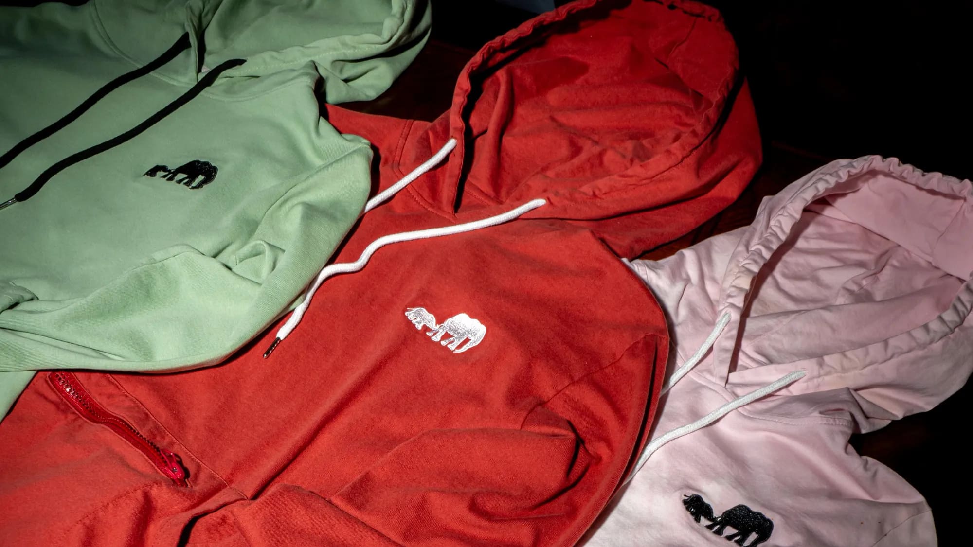 Sage, red and pale pink hoodies folded in a stack, hoods and drawcords overlapping