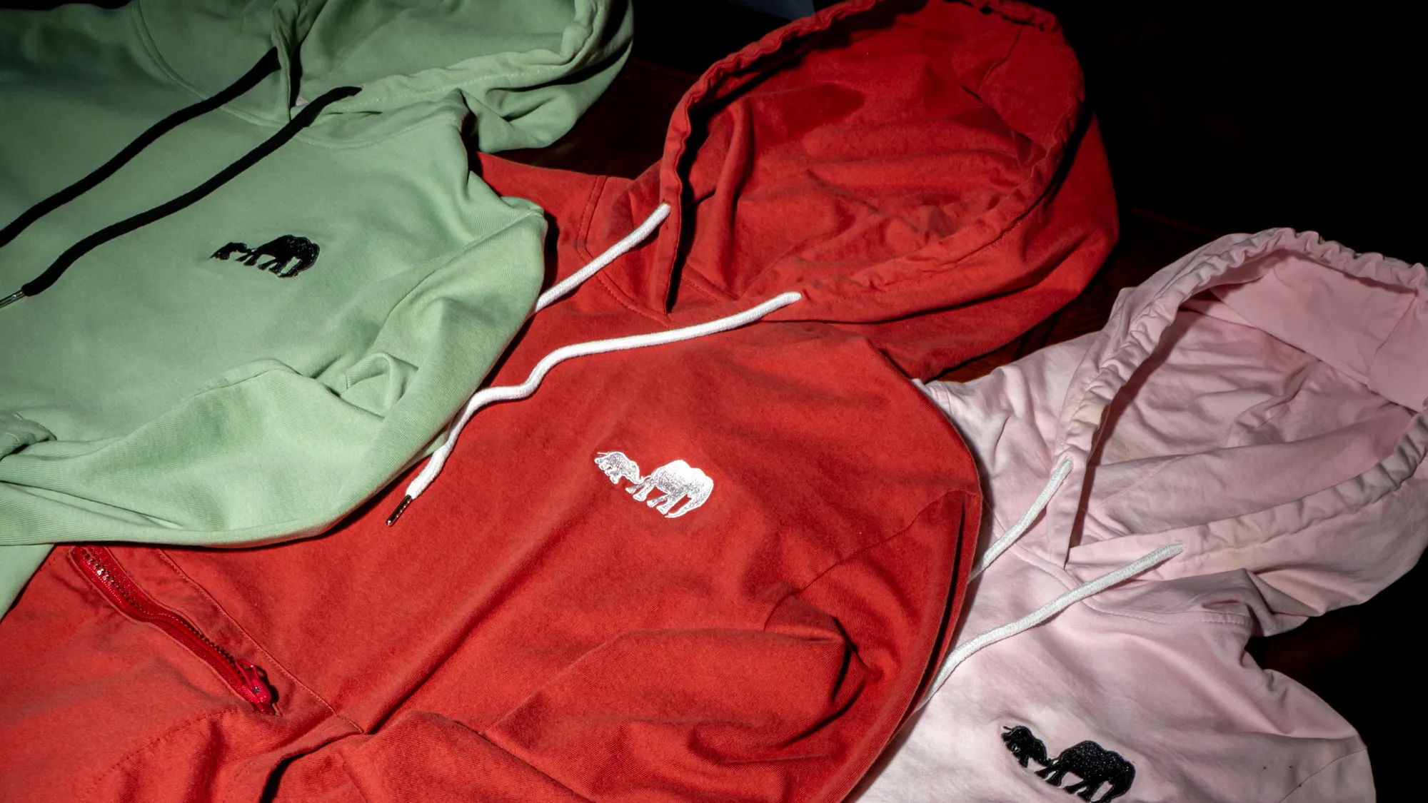 Sage, red and pale pink hoodies folded in a stack, hoods and drawcords overlapping