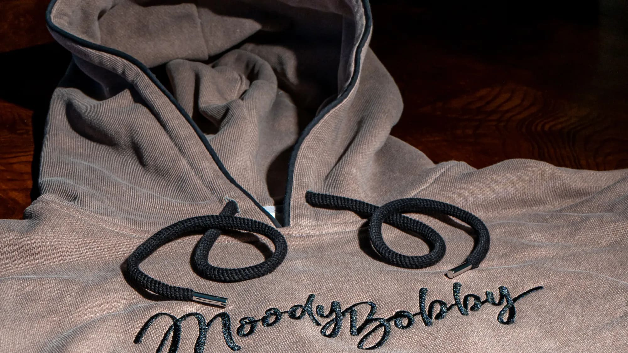 Close view of the embroidered Moody Bobby script on a grey hoodie, drawcords coiled across it
